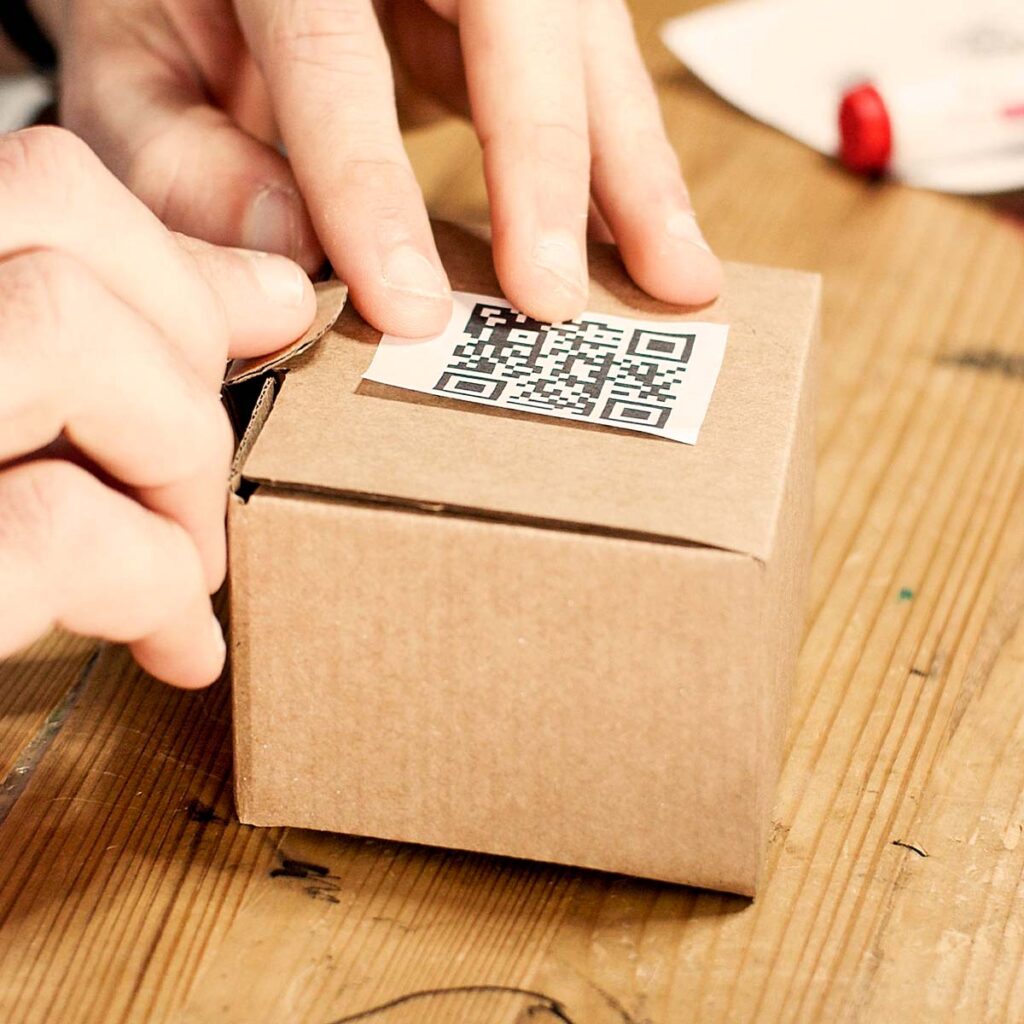 E-commerce store owner applying a custom generated QR code sticker to a cardboard shipping box to direct customers to a website URL for quick reordering.